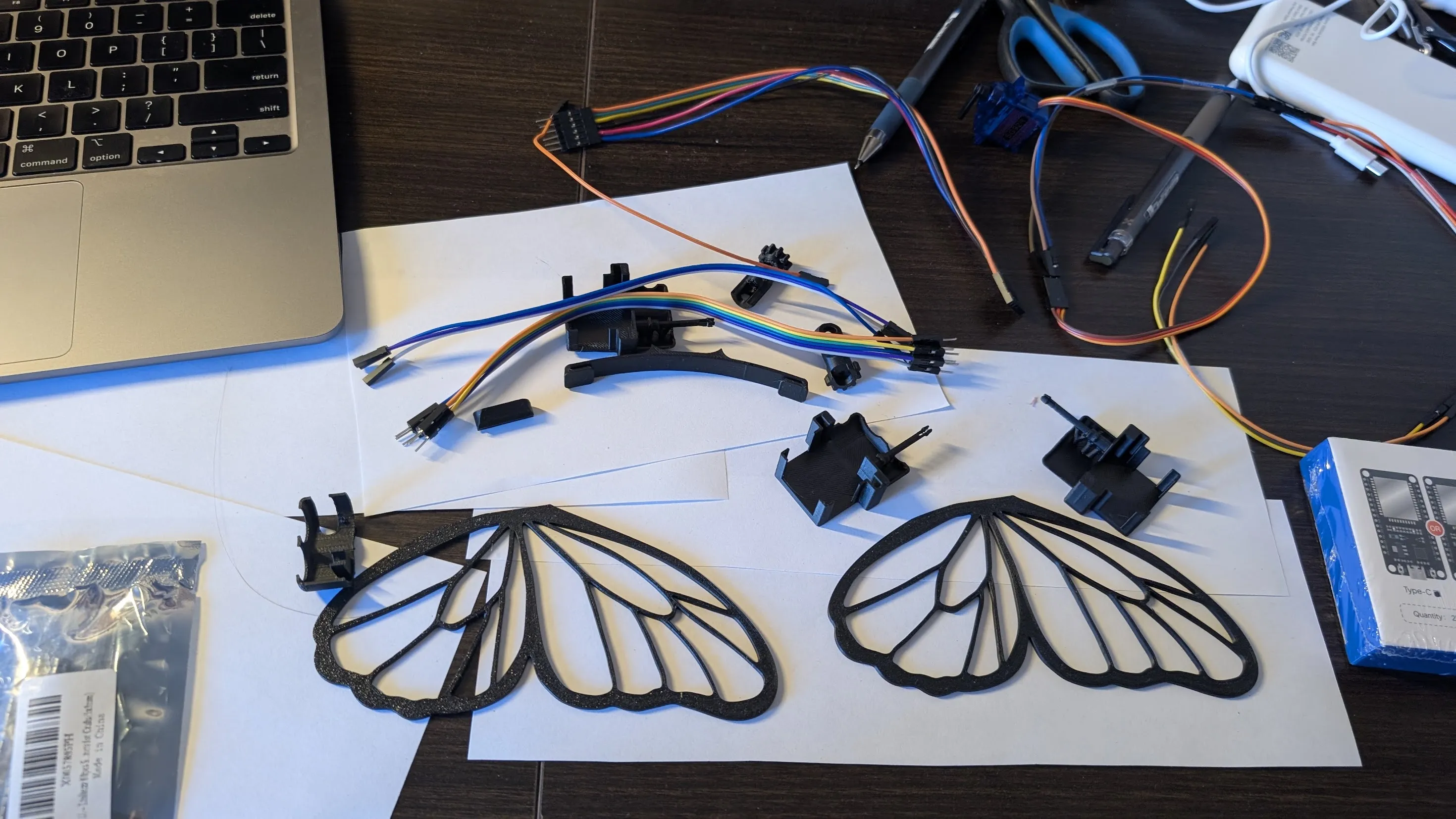 3d-printed butterfly wings and servo mounts laid out on a desk with jumper wires